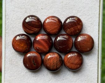 May include: Ten polished, round gemstones with a reddish-brown color and a glossy finish are arranged in a white tray. The stones have a chatoyant effect, displaying bands of light. These cabochons are suitable for jewelry making or collecting.