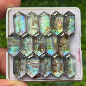 May include: A collection of fourteen hexagonal, faceted gemstones arranged in a grid on a white display tray. Each stone features a translucent, iridescent surface with swirling patterns of green, blue, gold, and silver, reflecting light.