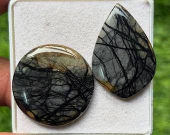 May include: Two polished gemstones, one round and one teardrop-shaped, displayed in a white tray. The stones feature a gray and tan base color with intricate black veining patterns. The gemstones are likely for jewelry making.