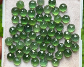 Op de afbeelding: Een verzameling gefacetteerde, ronde, groene edelstenen, gepresenteerd in een witte tray. De edelstenen hebben een gepolijst, reflecterend oppervlak en variëren enigszins in kleur. De tray wordt door een hand vastgehouden.