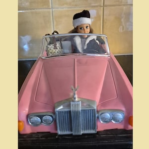 Puede incluir: Un coche descapotable de juguete rosa con una muñeca que lleva un sombrero blanco y negro y una chaqueta negra. El coche tiene una parrilla y faros plateados. Un pequeño bolso es visible en el coche.