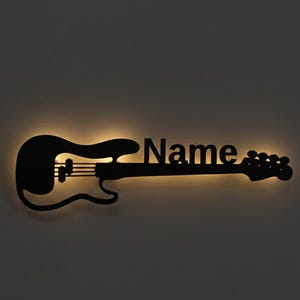 Peut inclure: Décoration murale noire en forme de guitare basse avec le mot "Name" découpé. La guitare a une lueur jaune et chaude émanant de derrière le motif découpé, créant une silhouette saisissante sur un fond sombre.