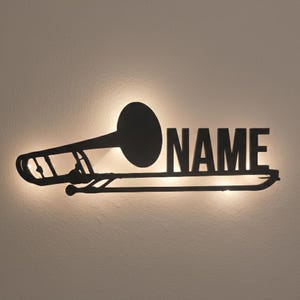 Puede incluir: Arte de pared de trombón de metal negro con la palabra "NAME" en letras de bloque. El trombón se muestra como una silueta contra una luz cálida y brillante, creando un contraste llamativo. Adecuado para una sala de música o como decoración única.
