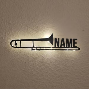 Puede incluir: Silueta de trombón de metal negro con la palabra "NAME" recortada. El trombón está iluminado con pequeñas luces blancas cálidas, creando un efecto brillante contra una pared texturizada. Decoración de pared de instrumento musical única.