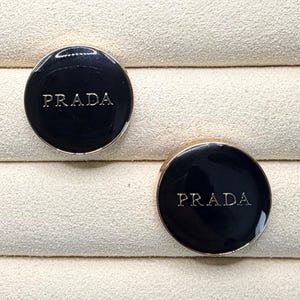 May include: Two black Prada cufflinks with silver lettering, displayed in a wooden jewelry box. The cufflinks are round with a glossy finish, set against a cream-colored interior.