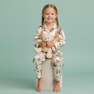 May include: A young girl wearing a cream-colored pajama set with a whimsical print of cartoon animals and apples. She is holding a beige teddy bear. The pajamas have a collared top and matching pants. The background is a solid light teal.