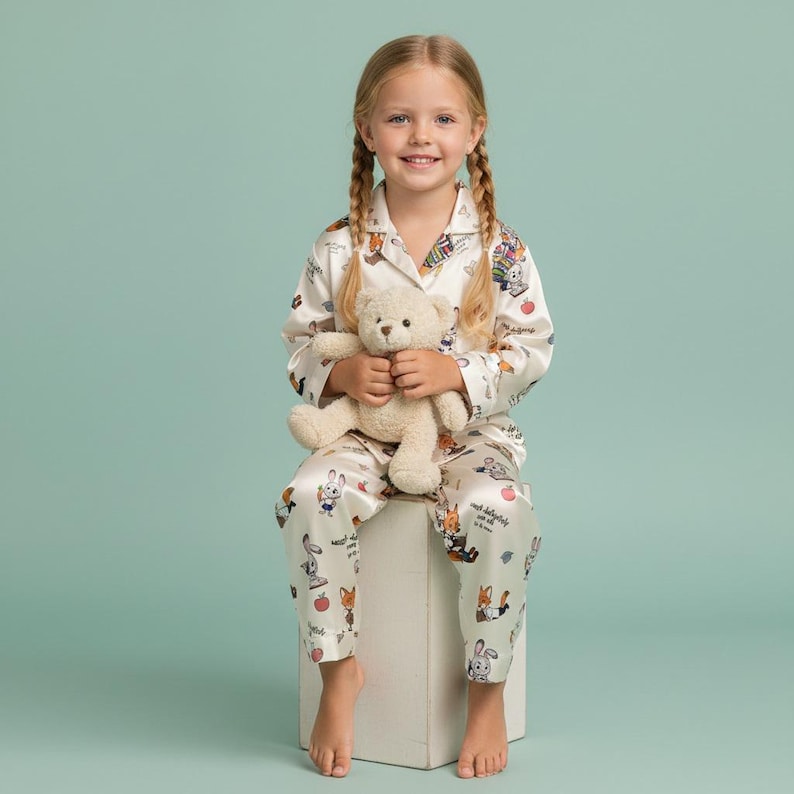 May include: A young girl wearing a cream-colored pajama set with a whimsical print of cartoon animals and apples. She is holding a beige teddy bear. The pajamas have a collared top and matching pants. The background is a solid light teal.