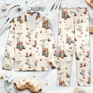 May include: Cream-colored pajama set with a repeating pattern of cartoon foxes and rabbits reading books. The set includes a button-down shirt and pants. The text "most definitely not all in a row" is printed on the fabric. A gold sleep mask is also visible.