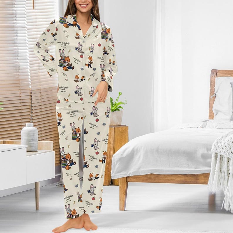 May include: Cream-colored pajama set featuring a repeating pattern of cartoon rabbits, foxes, and apples. The long-sleeved top has a collared neckline and button closure. The matching pants have a relaxed fit. The set is made of a soft, lightweight fabric.