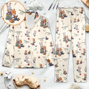 May include: Cream-colored pajama set featuring a repeating pattern of cartoon fox and rabbit characters reading books. The set includes a button-down shirt and pants. The text "most definitely not all in a row" is also visible.