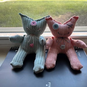 May include: Two handmade plush dolls crafted from ribbed fabric. The left doll is sage green with a pink button eye and a small pink heart button on its chest. The right doll is light pink with a blue button eye, a stitched eye, and a grey flower button.