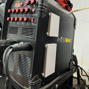 May include: A black PrimeWeld welding machine with red control knobs and a digital display. The machine has two white electrical outlets and a handle on top. A red cloth rests on the top of the machine.