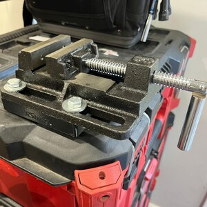 May include: A black metal bench vise with a silver screw and handle, mounted on a red and black toolbox. The vise has a sturdy design with a threaded rod for clamping objects. The vise is a tool used for woodworking and metalworking.