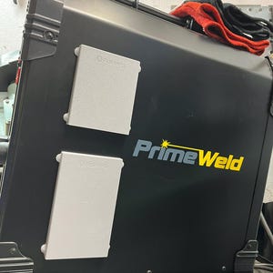 May include: A black PrimeWeld welding machine with two gray rectangular panels and the brand name in yellow and blue. A red and black cloth rests on top of the machine.