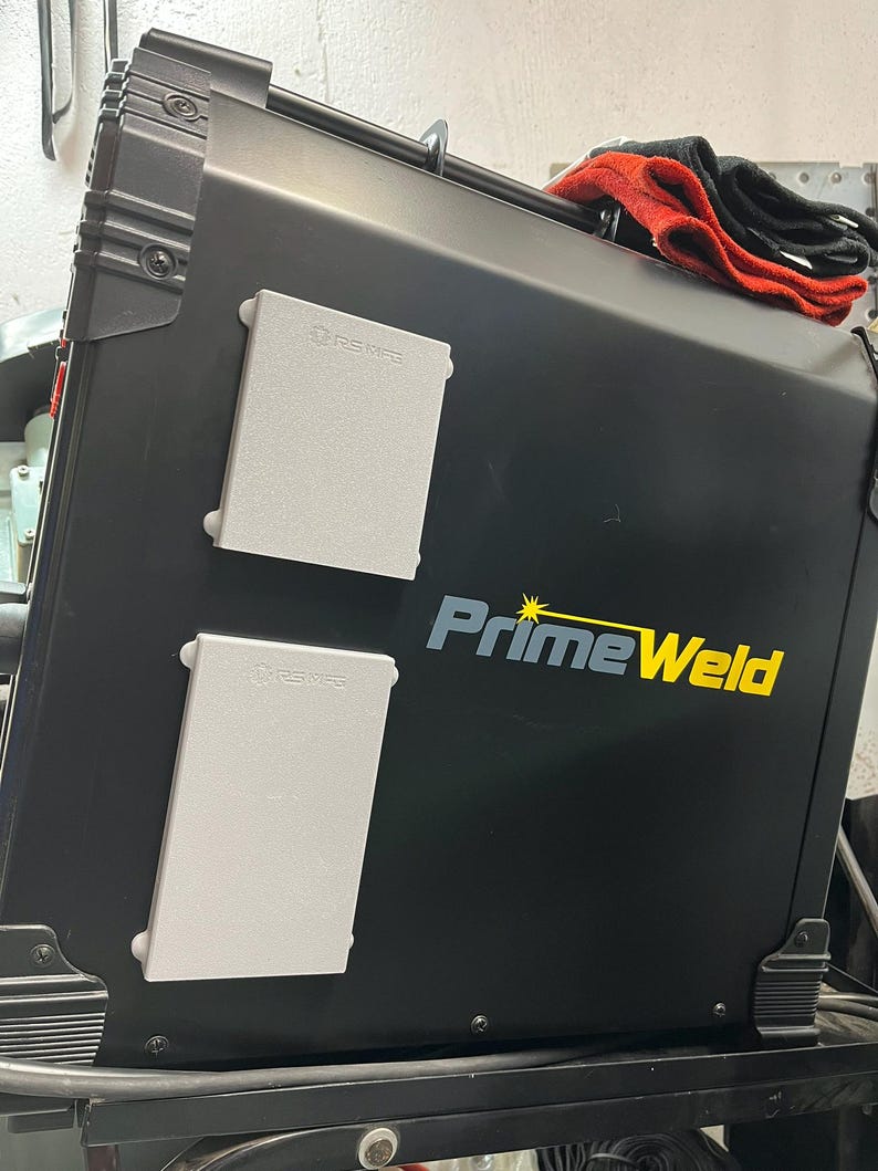 May include: A black PrimeWeld welding machine with two gray rectangular panels and the brand name in yellow and blue. A red and black cloth rests on top of the machine.