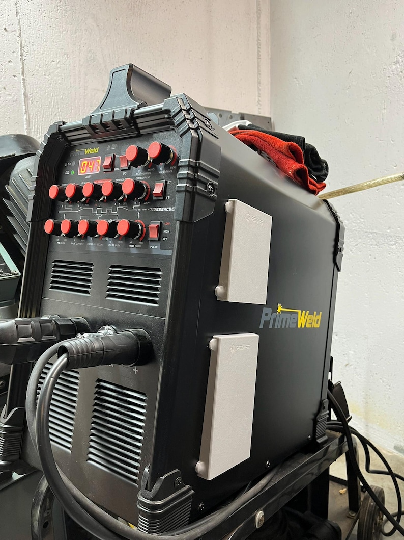 May include: A black PrimeWeld welding machine with red control knobs and a digital display. The machine has two white electrical outlets and a handle on top. A red cloth rests on the top of the machine.