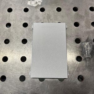 May include: A rectangular, silver-colored metal object with rounded corners and small, white, circular attachments on each corner. The text "RSM" is visible on the top surface. The object is on a metal surface with evenly spaced holes.
