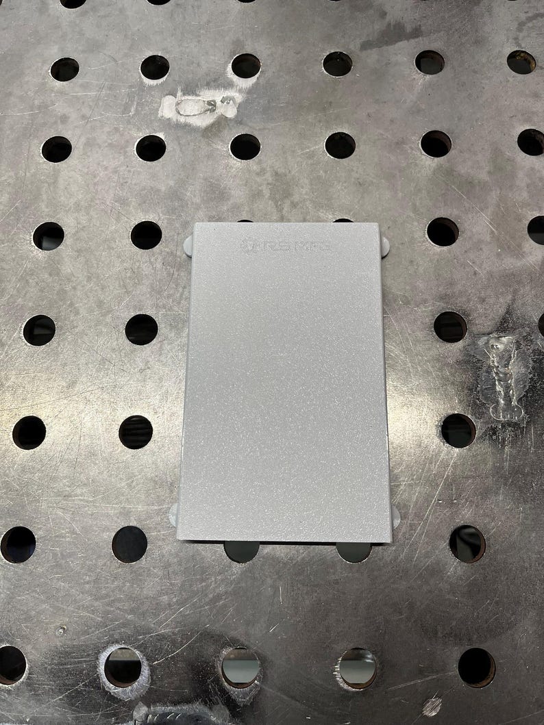 May include: A rectangular, silver-colored metal object with rounded corners and small, white, circular attachments on each corner. The text "RSM" is visible on the top surface. The object is on a metal surface with evenly spaced holes.