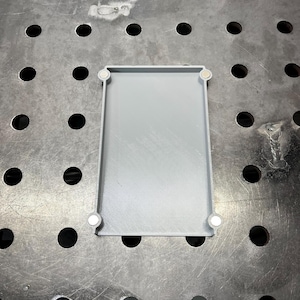 May include: A rectangular, light gray tray with rounded corners and a raised edge. Four small, white magnets are attached to the corners. The tray is sitting on a metal surface with evenly spaced holes.