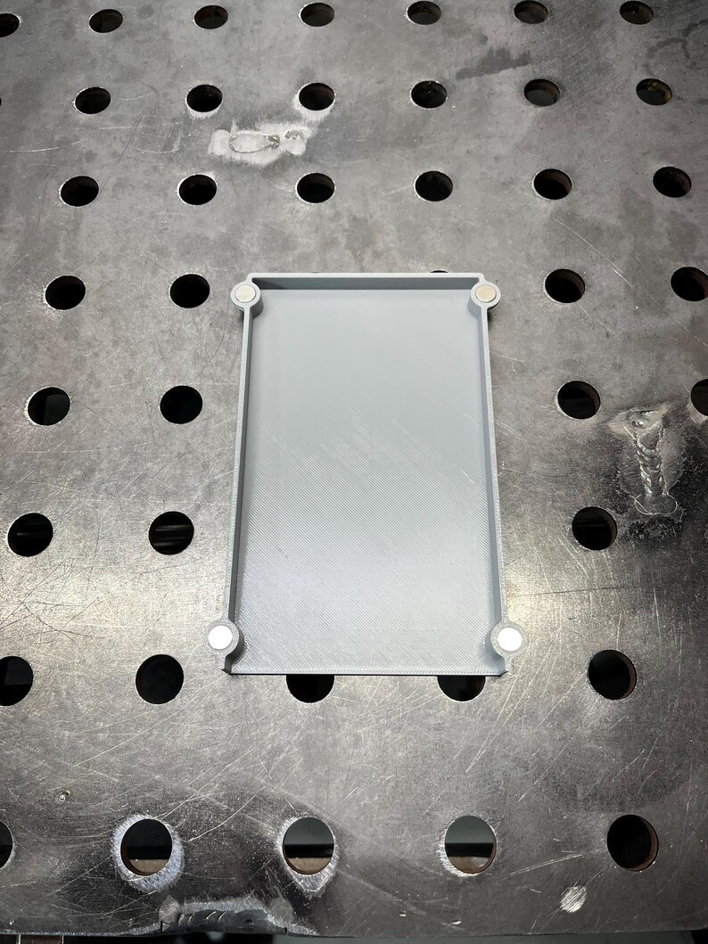 May include: A rectangular, light gray tray with rounded corners and a raised edge. Four small, white magnets are attached to the corners. The tray is sitting on a metal surface with evenly spaced holes.