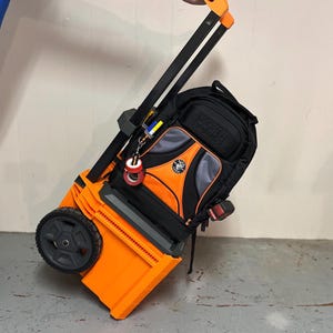 May include: An orange and black rolling tool bag with a black backpack attached. The bag has a telescopic handle and two wheels. The backpack features the "KLEIN TOOLS" logo and an orange and grey design.