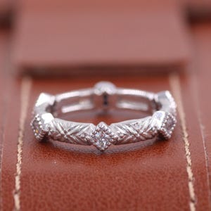 May include: A silver ring with a detailed geometric pattern and small, clear gemstones. The ring is set against a brown leather surface, highlighting its intricate design and craftsmanship. The ring's design features a series of diamond shapes.