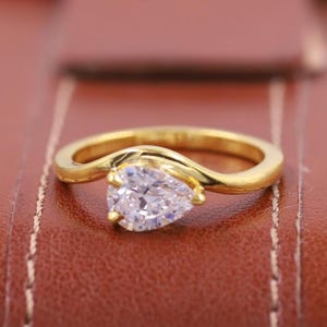 May include: A gold ring with a heart-shaped diamond. The ring is set on a brown leather surface. The ring's band is a simple, elegant design, and the diamond sparkles brightly. A beautiful piece of jewelry.