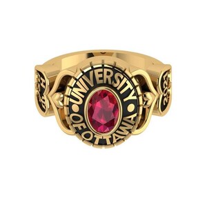 Puede incluir: Un anillo dorado con una piedra central de color rojo rubí de forma ovalada. El anillo lleva la inscripción "UNIVERSITY OF OTTAWA" en letras negras. El diseño incluye elementos decorativos y el año "2021".