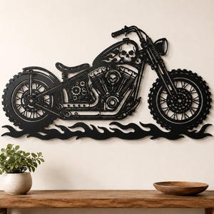 Op de afbeelding: Een zwart metalen wanddecoratie in de vorm van een motorfiets met een schedelontwerp op de tank en het motorblok. De motorfiets heeft gedetailleerde wielen, een stuur en een koplamp, rustend op een basis van gestileerde zwarte vlammen.