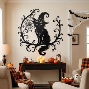 May include: A black metal wall art piece featuring a cat wearing a witch hat, positioned within a circular frame of swirling vines and a spider web. The design is flat, silhouette-style, and mounted on a light-colored wall above a wooden console table.