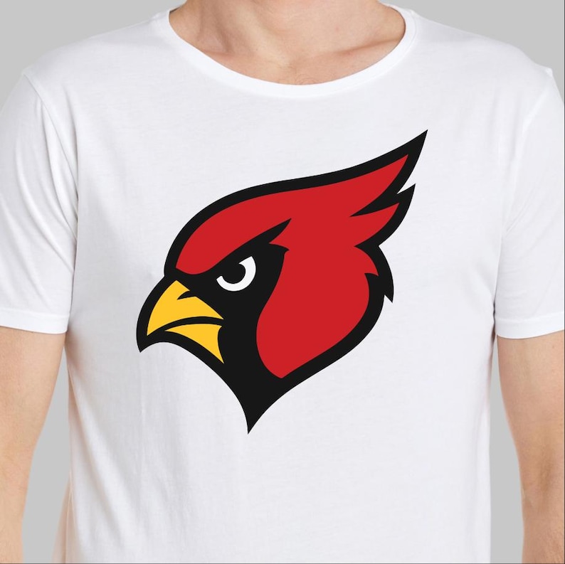 May include: White t-shirt featuring a stylized red cardinal head with a yellow beak and black accents. The cardinal graphic is centered on the chest. The shirt is made of a soft, white fabric.
