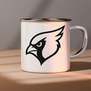 May include: White enamel mug with a silver rim and handle. The mug features a black graphic of a cardinal bird's head. The mug is sitting on a light brown surface.