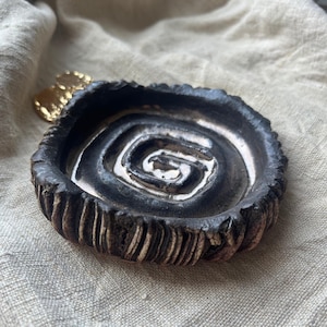 May include: A handmade ceramic dish with a dark, textured exterior and a spiral design in the center. The dish has a rough, organic shape and is likely used for holding small items. A small gold-colored decorative element is visible in the background.