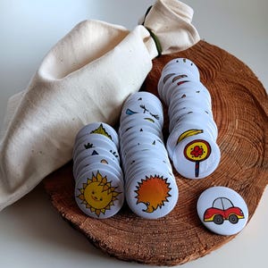 Może przedstawiać: Zestaw białych, ilustrowanych guzików z motywami słońca, jeża, samochodu i innych. Guziki ułożone są na drewnianym plastrze, obok beżowego worka ze sznurkiem.