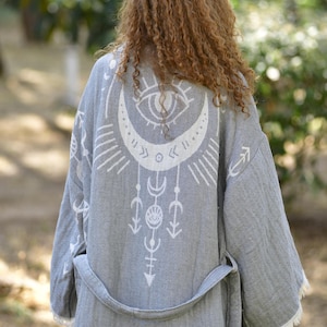 May include: A gray robe with a white celestial design on the back. The robe has a belt and fringe trim. The design includes an eye, crescent moon, and other geometric shapes. The robe is made of a soft, textured fabric.