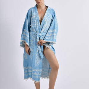 May include: A light blue patterned robe with a matching belt and fringed edges. The robe features a white geometric design and is made of a lightweight fabric. The model is wearing brown sandals.