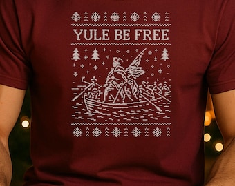 May include: Burgundy t-shirt with a white graphic design. The design features the text "YULE BE FREE" above a depiction of a boat with people and a flag, surrounded by snowflakes and trees. The shirt has short sleeves.