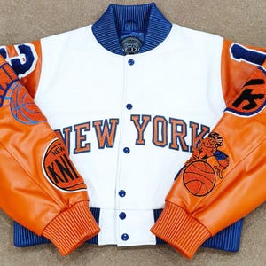 Peut inclure: Une veste universitaire blanche avec des manches orange, dotée de poignets et d'un col côtelés bleus. Le devant affiche le texte NEW YORK en lettres orange avec des contours bleus. Les manches sont ornées de patchs brodés de basket-ball.