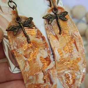 May include: A pair of earrings featuring a dragonfly charm and a marbled, peach-coloured resin base. The resin has a gold metallic finish.