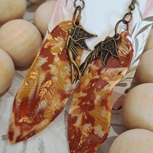 May include: A pair of dangle earrings with a brown and gold leaf design and a bronze fairy charm. The earrings are on a white background with a floral pattern.