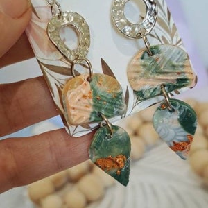 May include: A pair of earrings featuring a silver hammered circle charm and a green, peach, and gold abstract design. The earrings are handmade and have a unique, modern style.