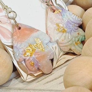 May include: A pair of handmade earrings with a unique, abstract design. The earrings are made of polymer clay and feature a mix of pastel colours, including pink, purple, and blue, with gold accents. The earrings are lightweight and dangle from silver hooks.