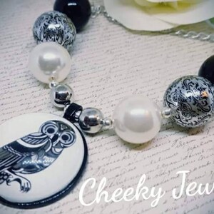 May include: A black and white owl charm necklace with white and black patterned beads. The necklace is on a white surface with the text "Cheeky Jewellery".