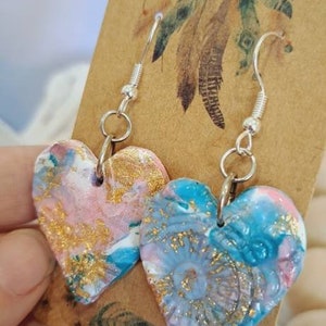 May include: A pair of heart-shaped earrings with a pink, blue and gold marbled design. The earrings are hanging from silver hooks.