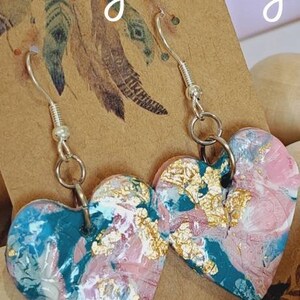 May include: A pair of heart-shaped earrings with a teal, pink and gold marbled design. The earrings are made of polymer clay and have silver hooks.