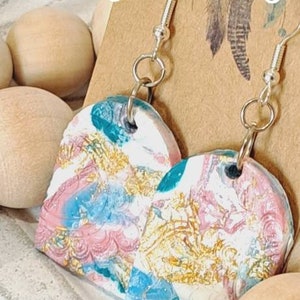 May include: A pair of heart-shaped earrings with a white, pink, blue and gold marbled design. The earrings are made of polymer clay and have silver hooks.