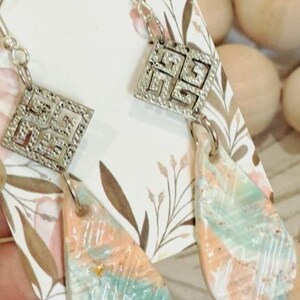 May include: A pair of dangle earrings featuring a silver geometric design and a light blue and peach coloured teardrop shaped pendant. The earrings are displayed on a white background with a floral pattern.