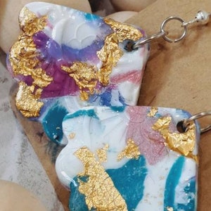 May include: A pair of colourful resin earrings with gold leaf accents. The earrings are shaped like abstract flowers and have a white, pink, blue, and teal colour scheme.