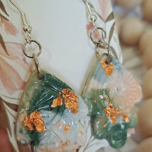 May include: A pair of earrings with a green, white and gold abstract design. The earrings are made of resin and have silver hooks.