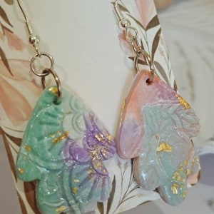 May include: A pair of handmade earrings featuring a pastel green and purple leaf design with gold accents. The earrings are on a white card with the text "Cheeky Jewellery".